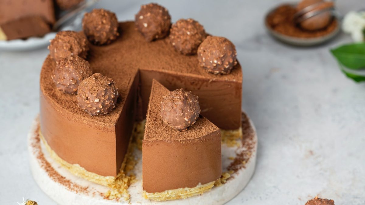 Grauer Steinuntergrund. Im Bildzentrum eine angeschnittene Schokoladentorte mit Rocher-Trüffeln.