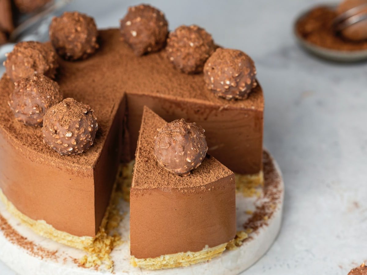 Grauer Steinuntergrund. Im Bildzentrum eine angeschnittene Schokoladentorte mit Rocher-Trüffeln.