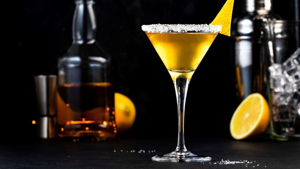 Sidecar Cocktail in Cocktailschale mit Zitronenzeste vor schwarzem Hintergrund. Im Hintergrund außerdem eine Flasche Cognac, Barmaß, Zitronen und Shaker. Frontalansicht.