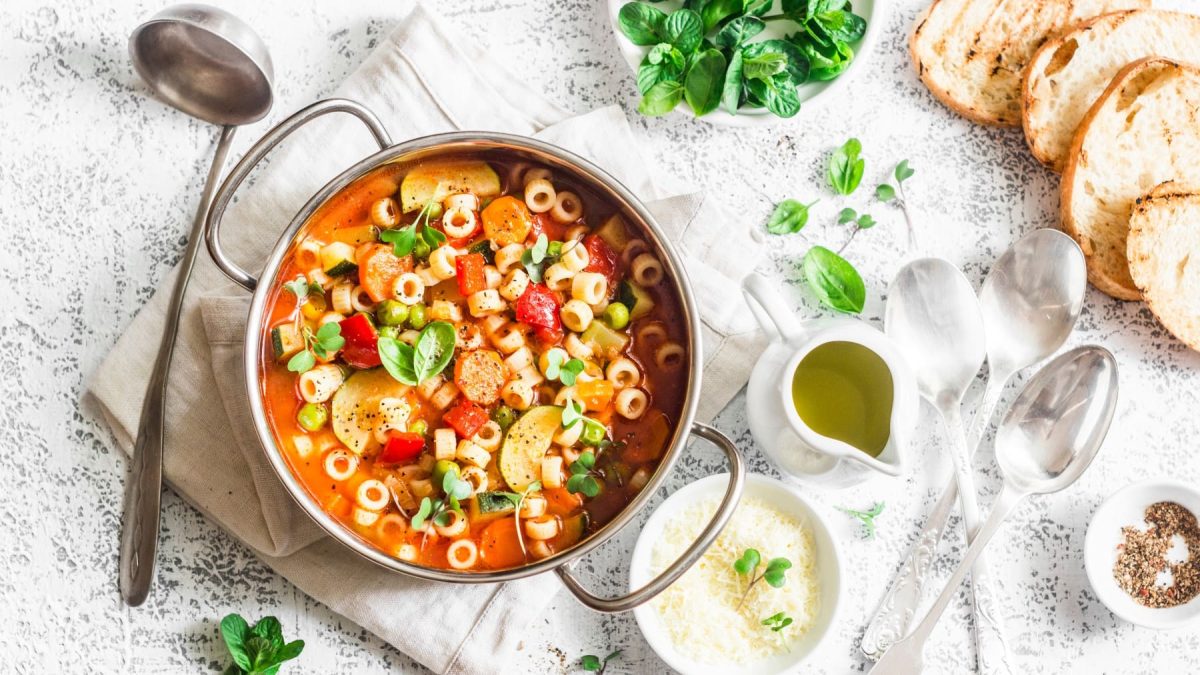 Sommerliche Minestrone: ein Topf mit Suppe, darin Nudeln und Gemüse, drumherum allerlei Zutaten.