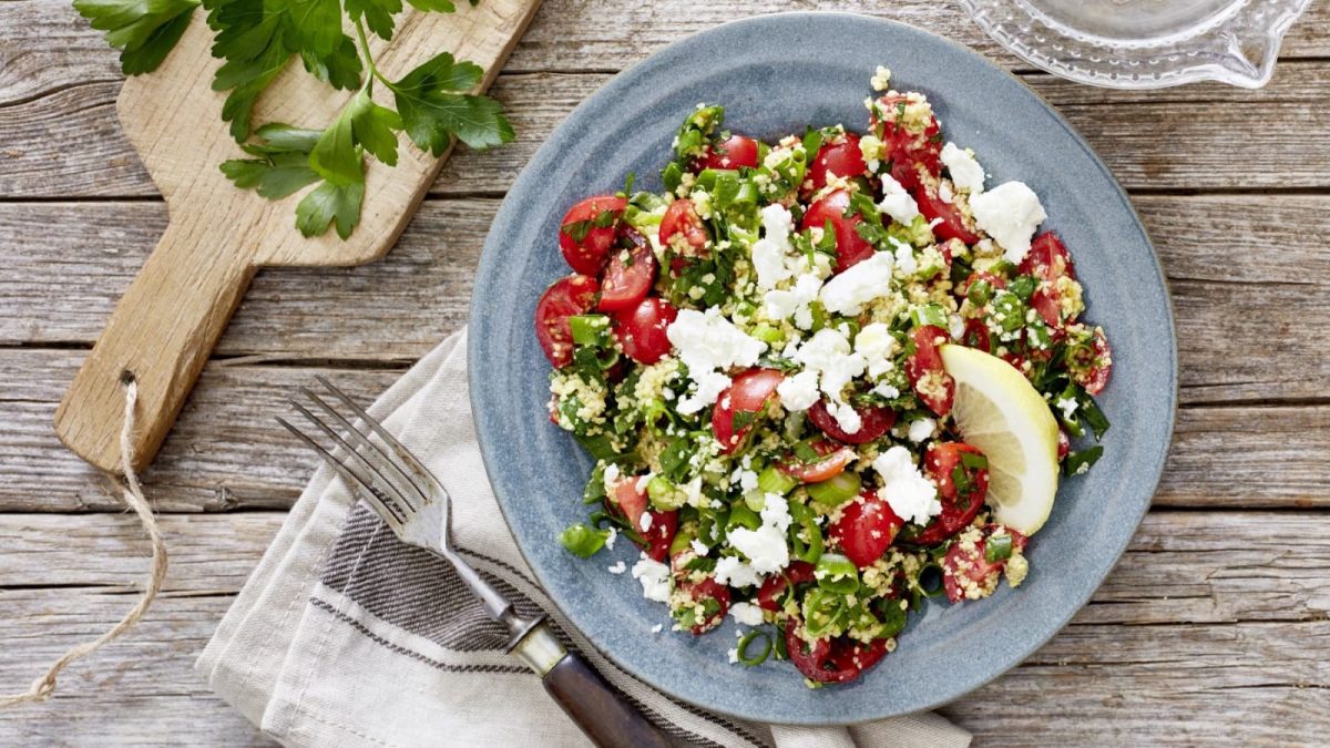 Draufsicht: Holzuntergrund mit einem Holzbrett oben links, einer beigen Stoffservierte unten und einem blauen Teller im Bildzentrum. Darauf ein Tomaten-Amaranth-Salat mit Petersilie.
