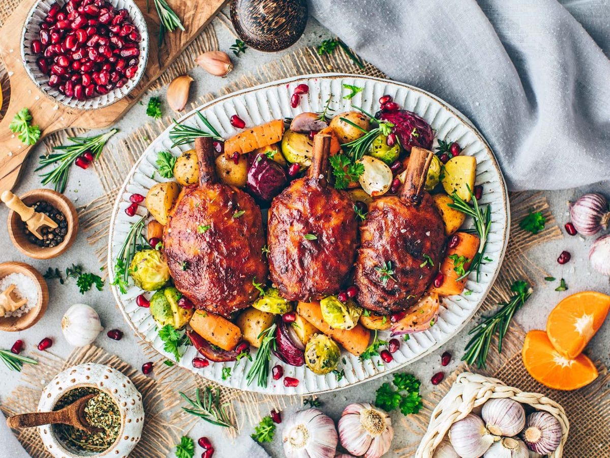 Ein weißer Teller mit den veganne Hähnchenkeulen auf Ofengemüse. Drum herum zahlreiche Dekoration in Form von Gewürzen.