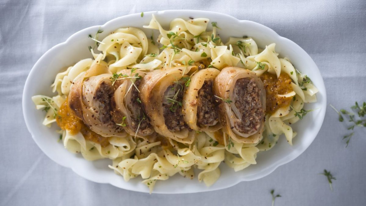 Draufsicht: Weiße Stofftischdecke. Im Bildzentrum eine ovale weiße Servierplatte auf der vegane Kohlrouladen-Scheiben auf einem Nudelbett liegen. Garniert mit frischen Kräutern.
