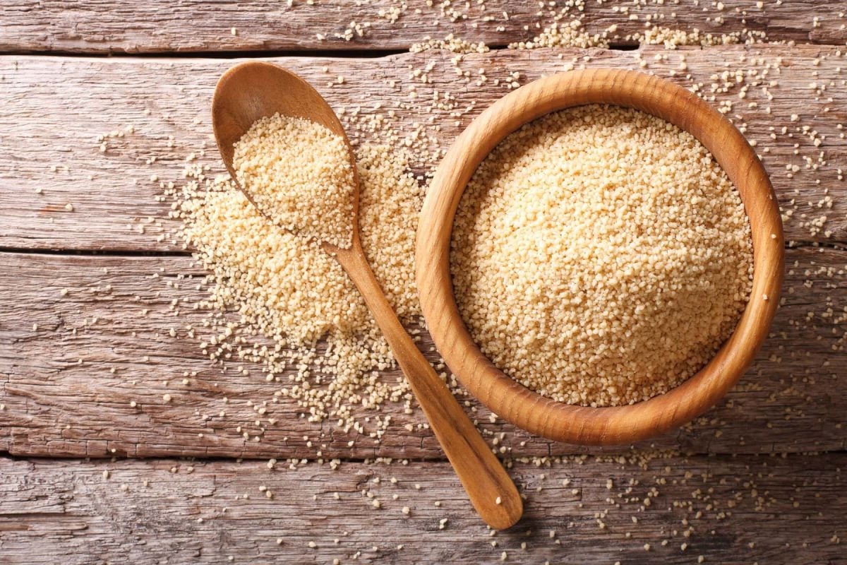 Draufsicht: Untergrund aus Holz. Darauf eine runde Schale mit Couscous sowie in Löffel aus Holz mit Couscous.