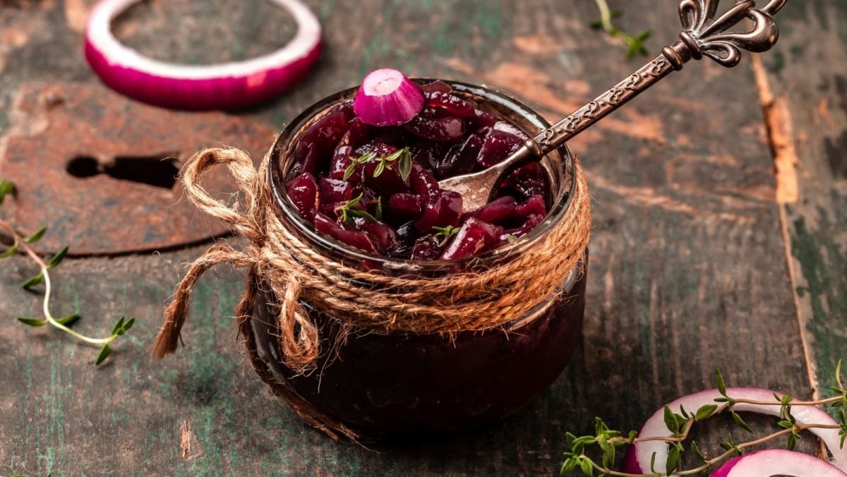 Schräge Ansicht: Holztisch auf dem ein kleines Schraubglas steht. Darin enthalten würziges Zwiebel-Chutney aus roten Zwiebeln, mit Thymian.