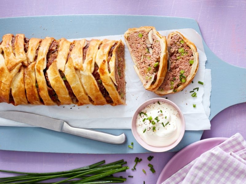 Hackbraten im Blätterteigmantel, angeschnitten serviert auf einem Holzbrett mit Joghurt-Sauce dazu.