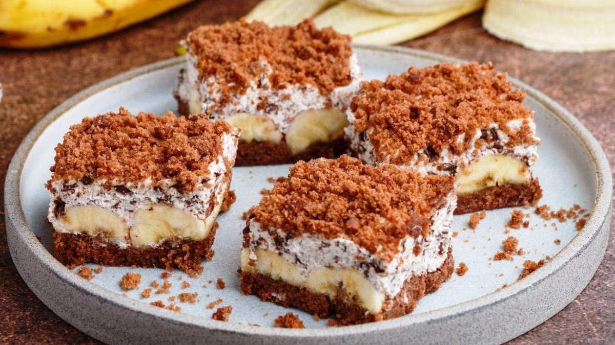Vier Stücken von dem Bananenkuchen auf einem dunkelbraunem Tisch. Im Hintergrund eine geschälte Banane und ein paar Schokostücken.