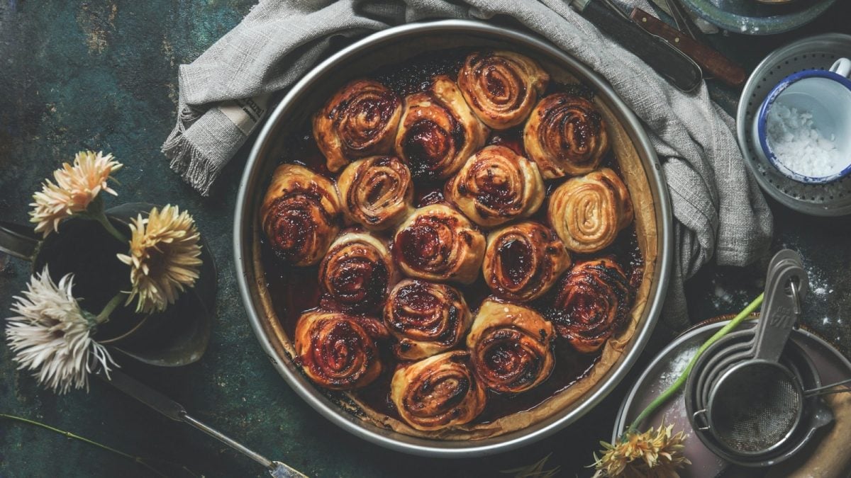 Draufsicht: Dunkler Untergrund auf dem mittig eine Backform mit buttrigen Blaubeermarmeladenschnecken steht. Darum verteilt ein paar Backutensilien und Blumen.