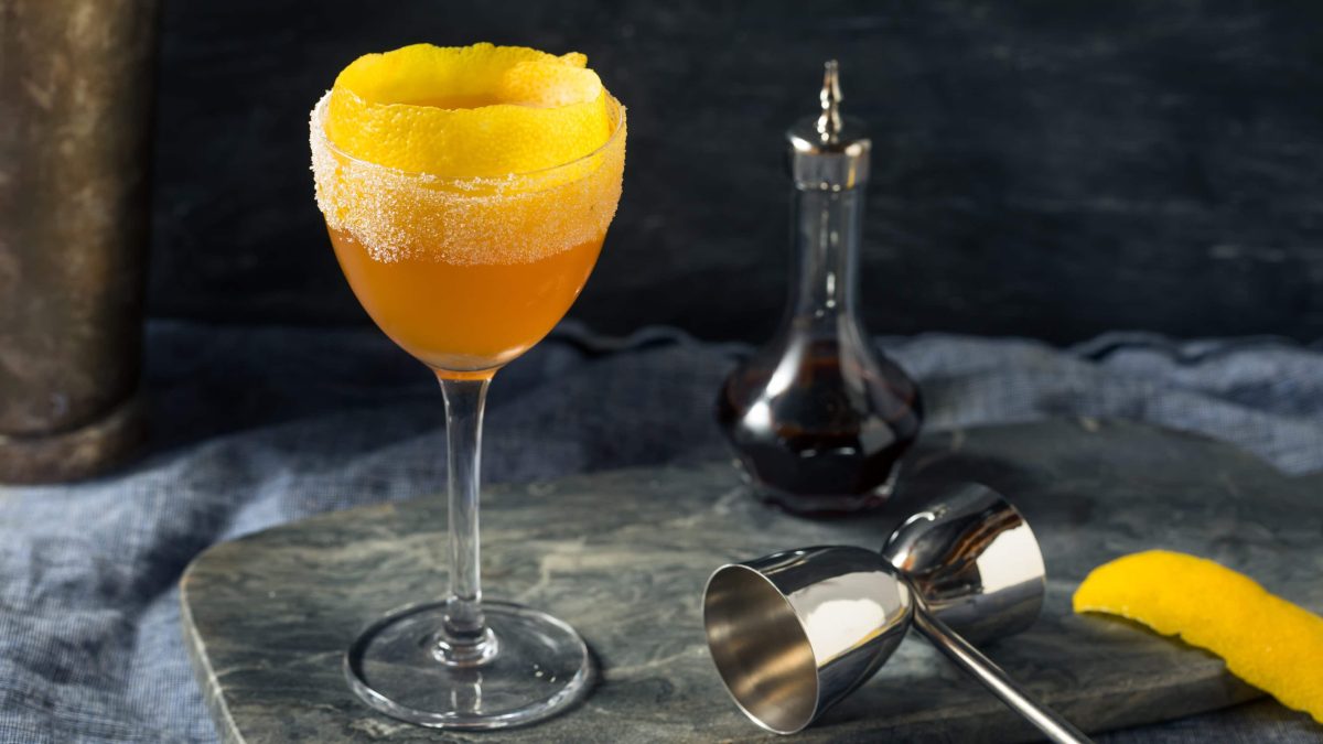 Brandy Crusta auf Stein mit Barmaß, Zitronenzeste und Karaffe mit rotem Likör. Frontalansicht.