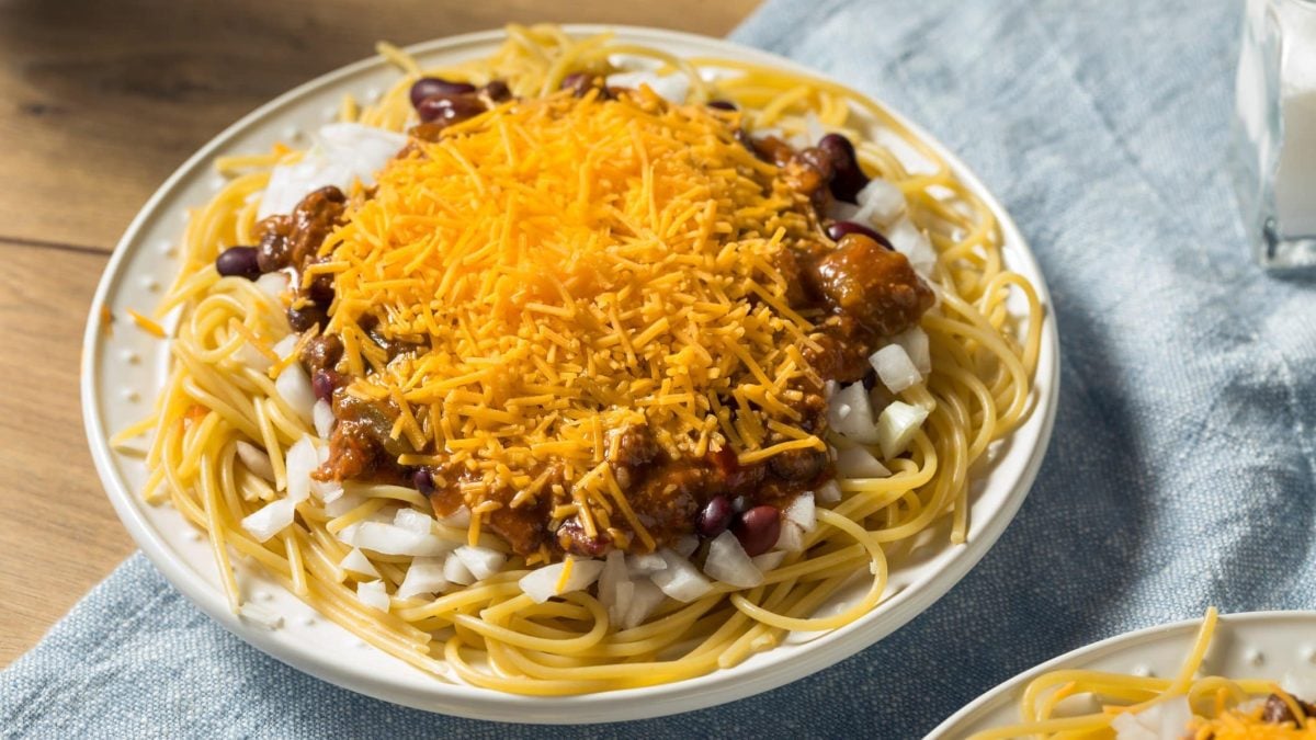 Cincinnati Chili mit Cheddar auf Pastateller, blauem Tuch und Holz. Unten eine weitere Portion angedeutet. Draufsicht.