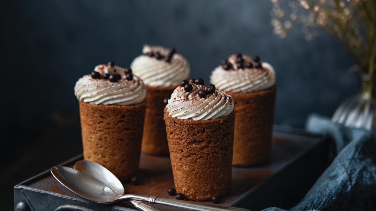 Dunkler Hintergrund. Vier Cookie-Cups nach Tiramisu-Art auf einem dunklen Koffer. Davor zwei Silberlöffel. Im rechten Bildrand eine blaue Stoffservierte und im Hintergrund Schleiherkraut.