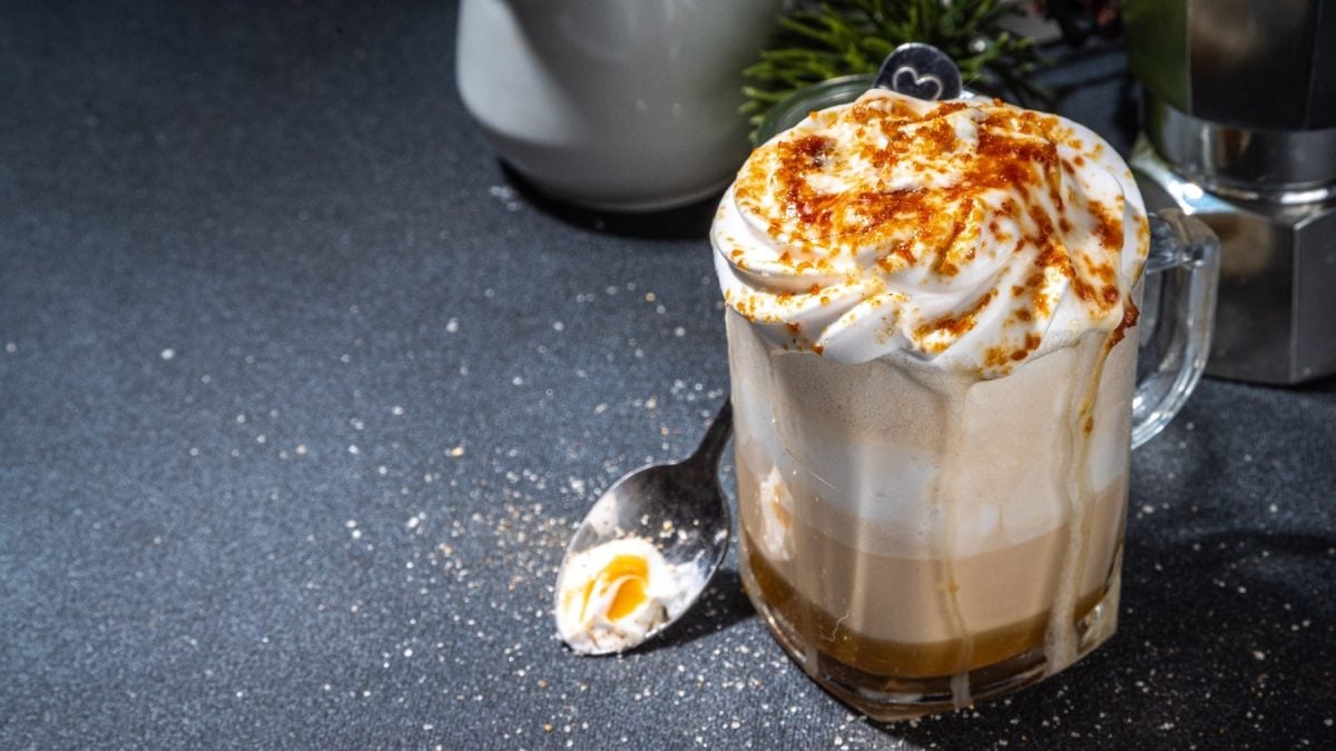 Eine Tasse mit dem Crème-Brûlée-Latte auf einem dunklen Untergrund. Im Hintergrund unscharf Kaffeeequipment.