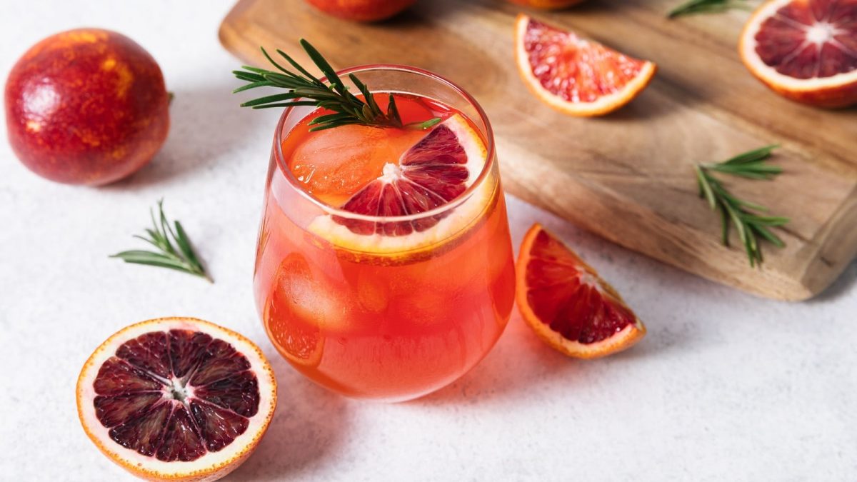Ein Glas mit Blutorangen Whisky Sour neben einem Holzbrett und frischen Blutorangenscheiben.