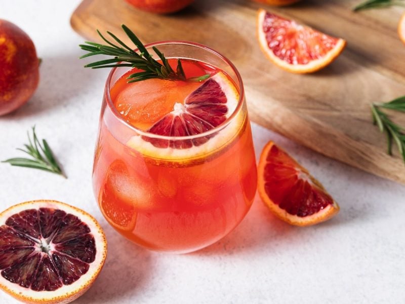 Ein Glas mit Blutorangen Whisky Sour neben einem Holzbrett und frischen Blutorangenscheiben.