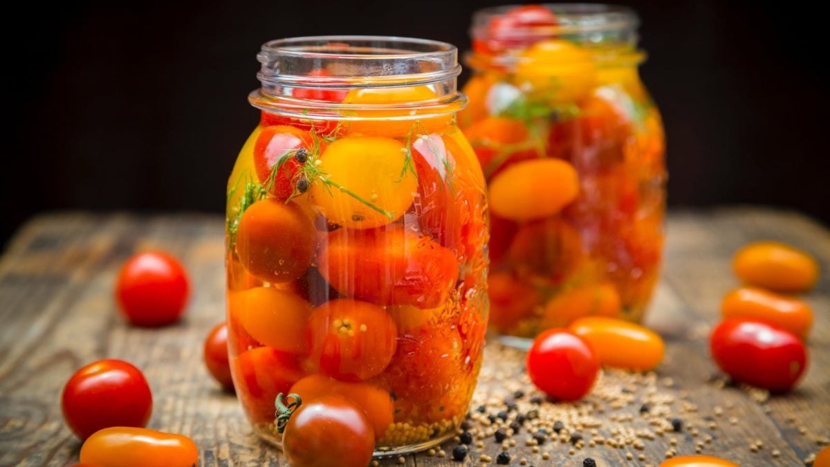 Nahaufnahme: Schwarzer Hintergrund, mit Holztisch auf dem zwei Schraubgläser mit eingelegten Tomaten in Honig-Sud stehen. Drumherum ein paar frische Tomaten.