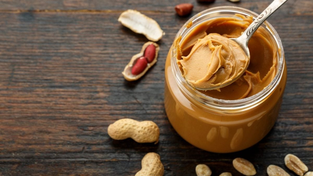 Draufsicht: Auf einem dunklen Untergrund steht ein Glas Edrnussbutter. In dem Glas ist ein Löffel. Drum herum liegen ganze Erdnüsse und Erdnusschalen.