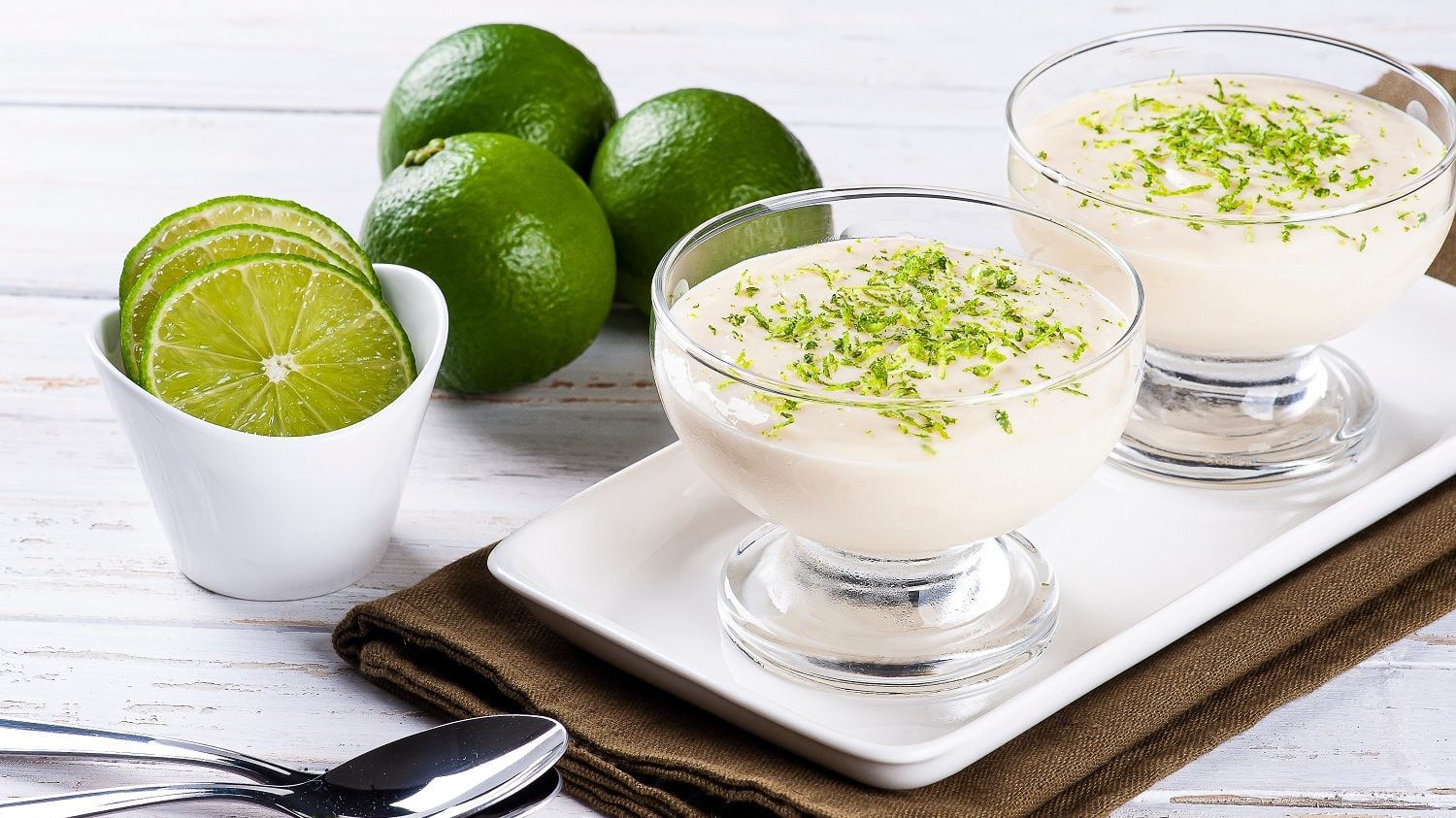 Frontalsicht: Auf einem weißen Holztisch liegt ein dunkles Holzbrettchen. Darauf sind zwei Gläser mit einer Frischkäse-Mousse. Drumherum liegen zwei Löffel sowie einige Limetten.