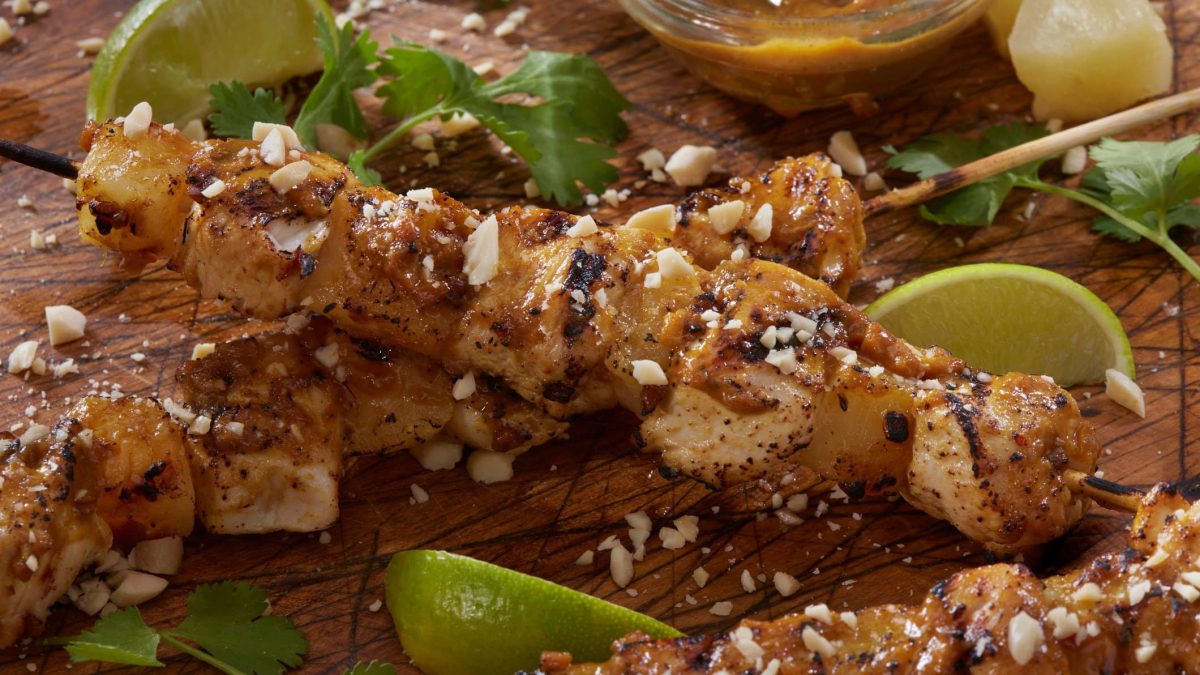 Gegrillte Ananas-Hähnchenspieße mit Erdnussglasur mit gehackten Erdnüssen, Limettenstücken und Koriander auf Holz. Draufsicht.