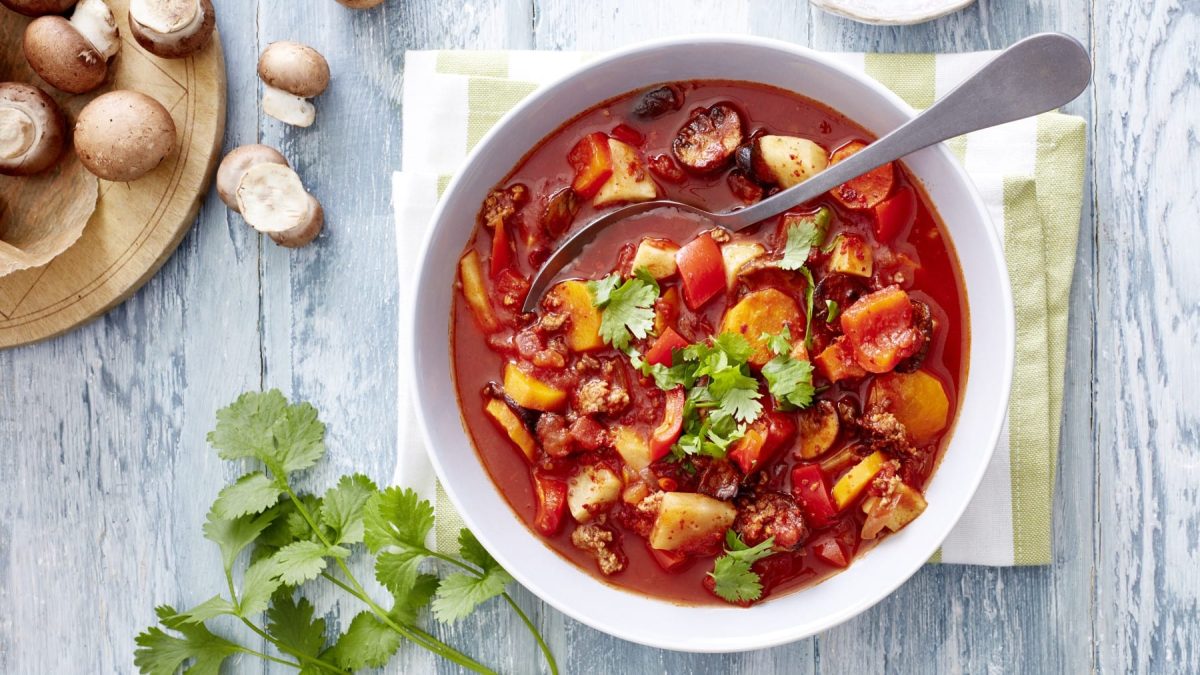 Gemüseeintopf mit Hack und Chili und Koriander in der Draufsicht, daneben Pilze, alles auf hellem Holzhintergrund.