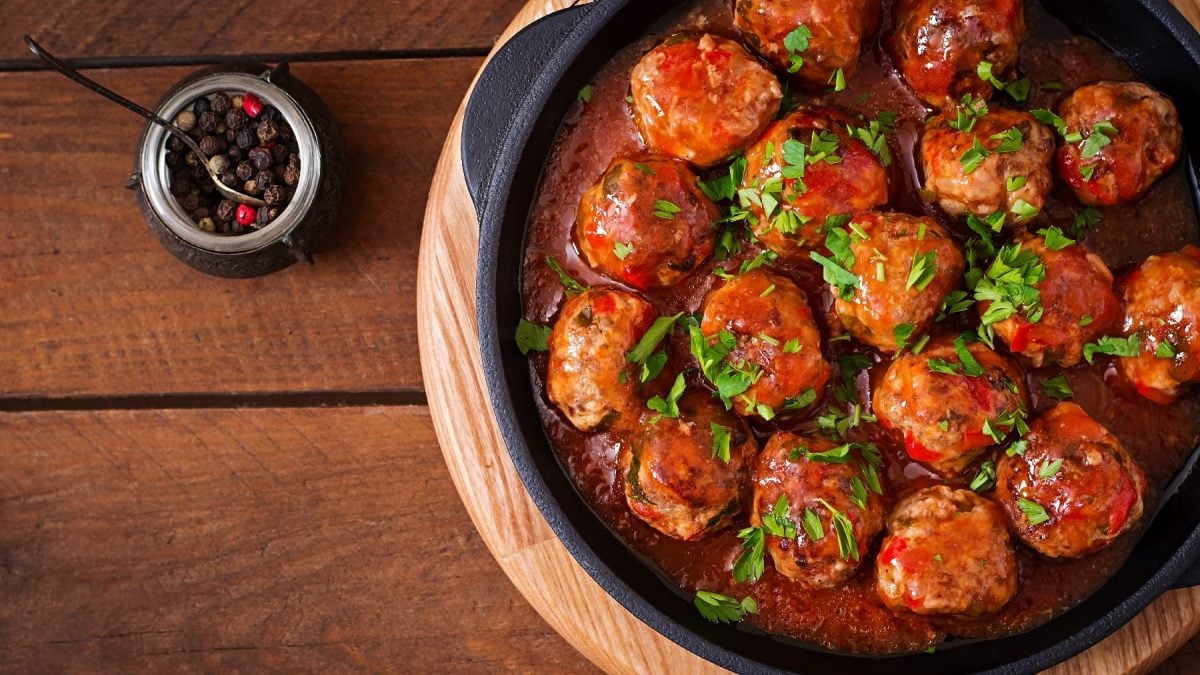 Eine gusseiserne Pfanne mit den Hackbällchen in Sweet-Chili-Sauce auf einem dunklen Holztisch von oben fotografiert.