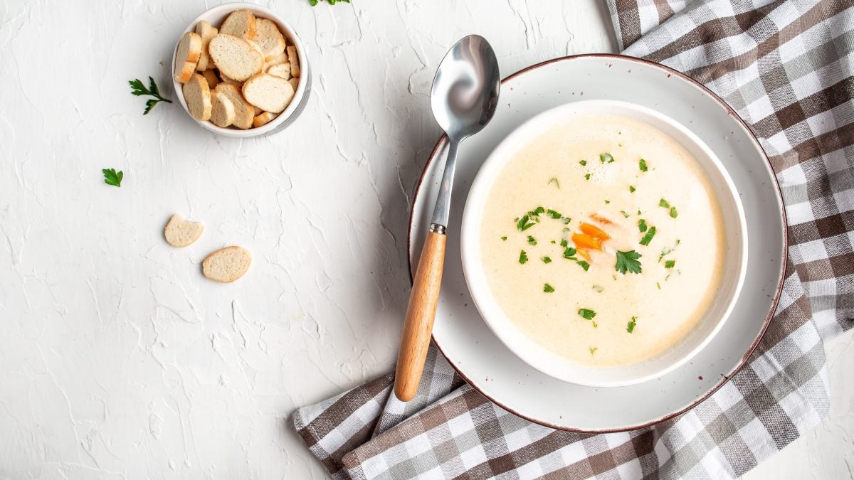 Draufsicht: eine Feine Hähnchen-Cremesuppe sowie ein Teller mit Croûtons und ein kariertes Küchentuch stehen auf einer hellen Tischplatte.