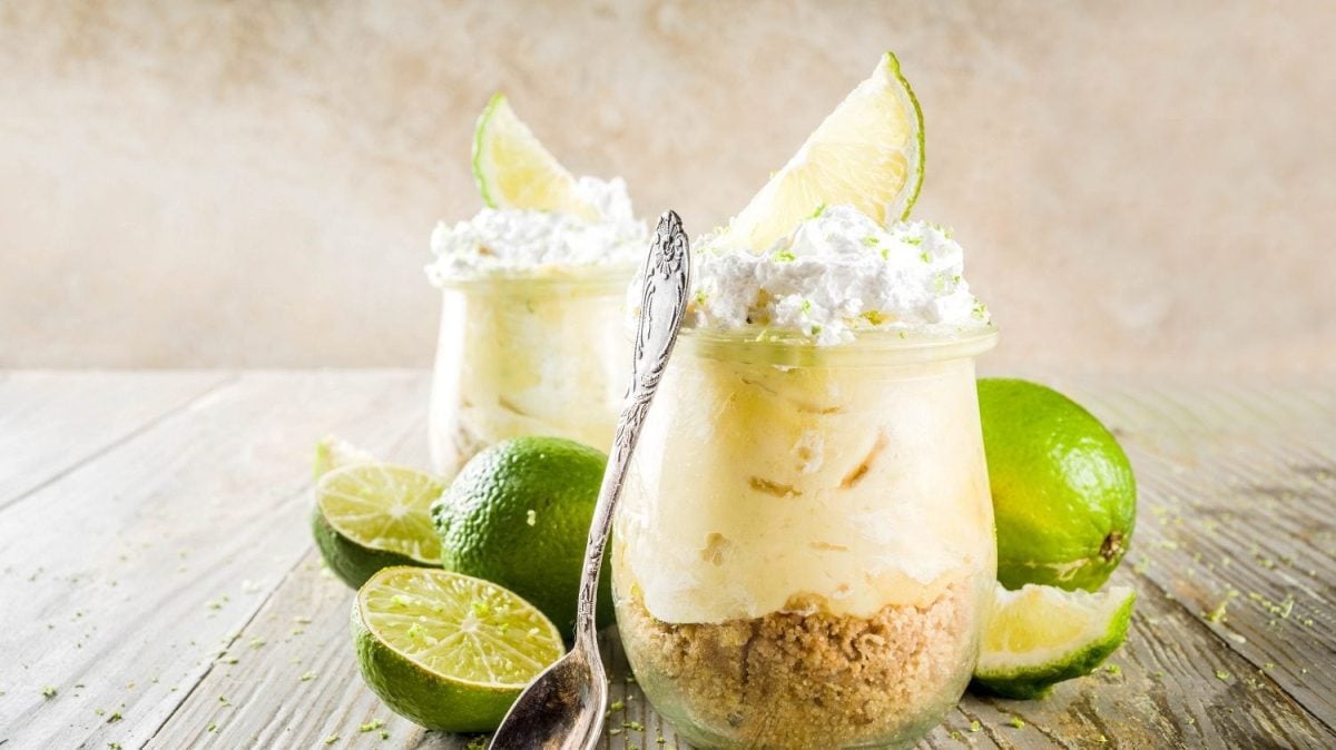 Frontalsicht: Zwei Kuchen im Glas, beziehungsweise Käse-Limetten-Kuchen, stehen auf einem Holztisch.. Drum herum liegen Limetten.