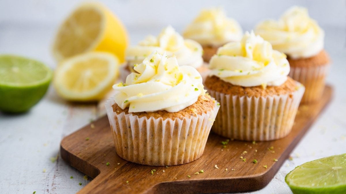 Ein paar Kokos-Limetten-Cupcakes auf einem Holzbrett, drumherum halbierte Limetten und Zitronen.