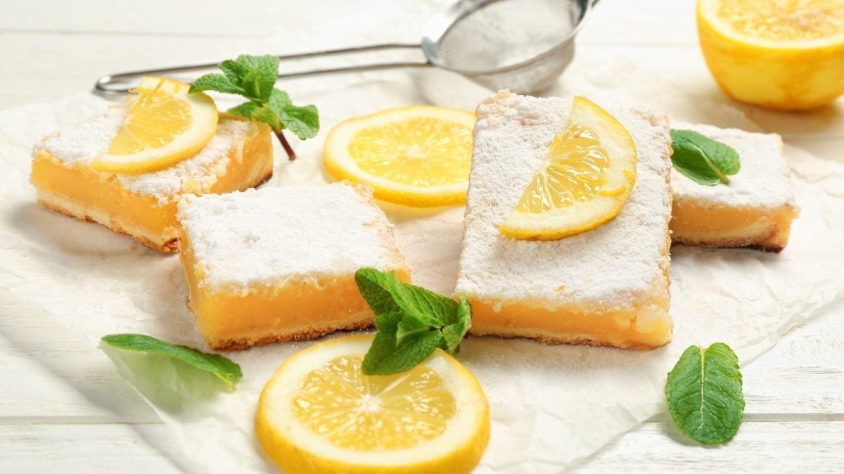 Frontalsicht: auf einem weißen Untergrund aus Holz liegen mehrere Lemon Bars bzw. Zitronenschnitten, Zitronenscheiben und Minzbätter.