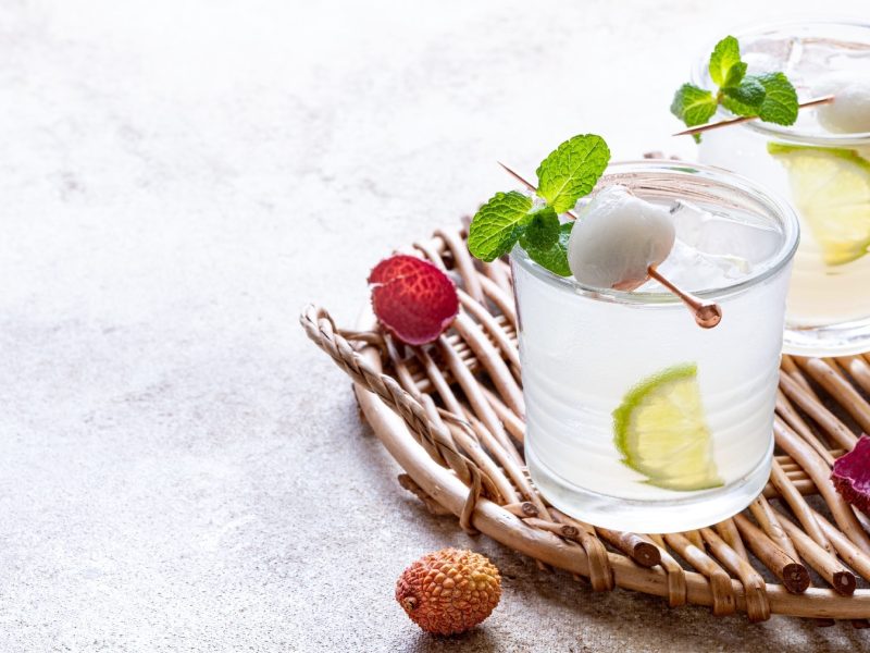 Zwei Gläser mit Litschi-Mojito auf einem hellgrauen Tisch mit frischen Litschis von schräg oben fotografiert.