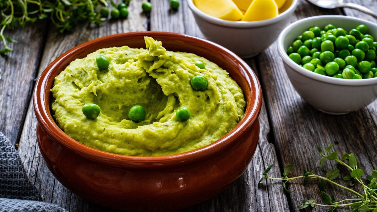Frontalsicht: Dunkler Untergrund aus Holz. Eine Tonform mit Mushy Potatoe Peas steht auf darauf. Drum herum ist eine schale mit Erbsen, eine Schale mit Kartoffeln und Minze.