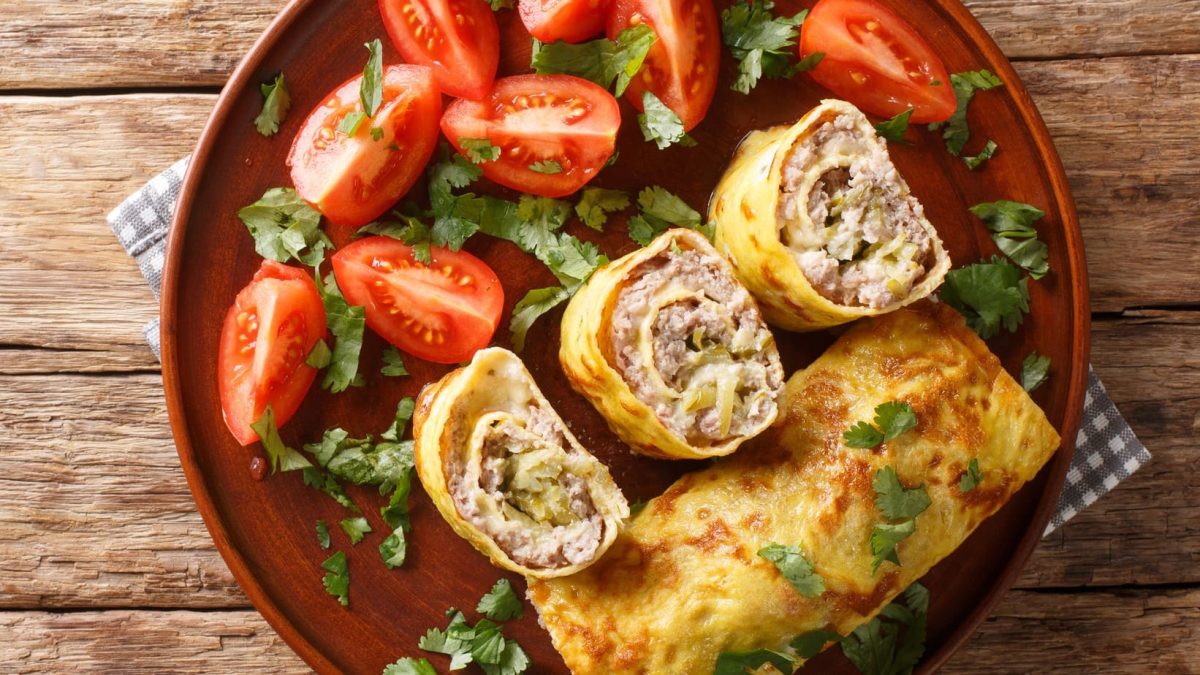 Angeschnittene Omelett-Röllchen mit Hackfleisch-Gurken-Käse-Füllung. Daneben Tomaten.