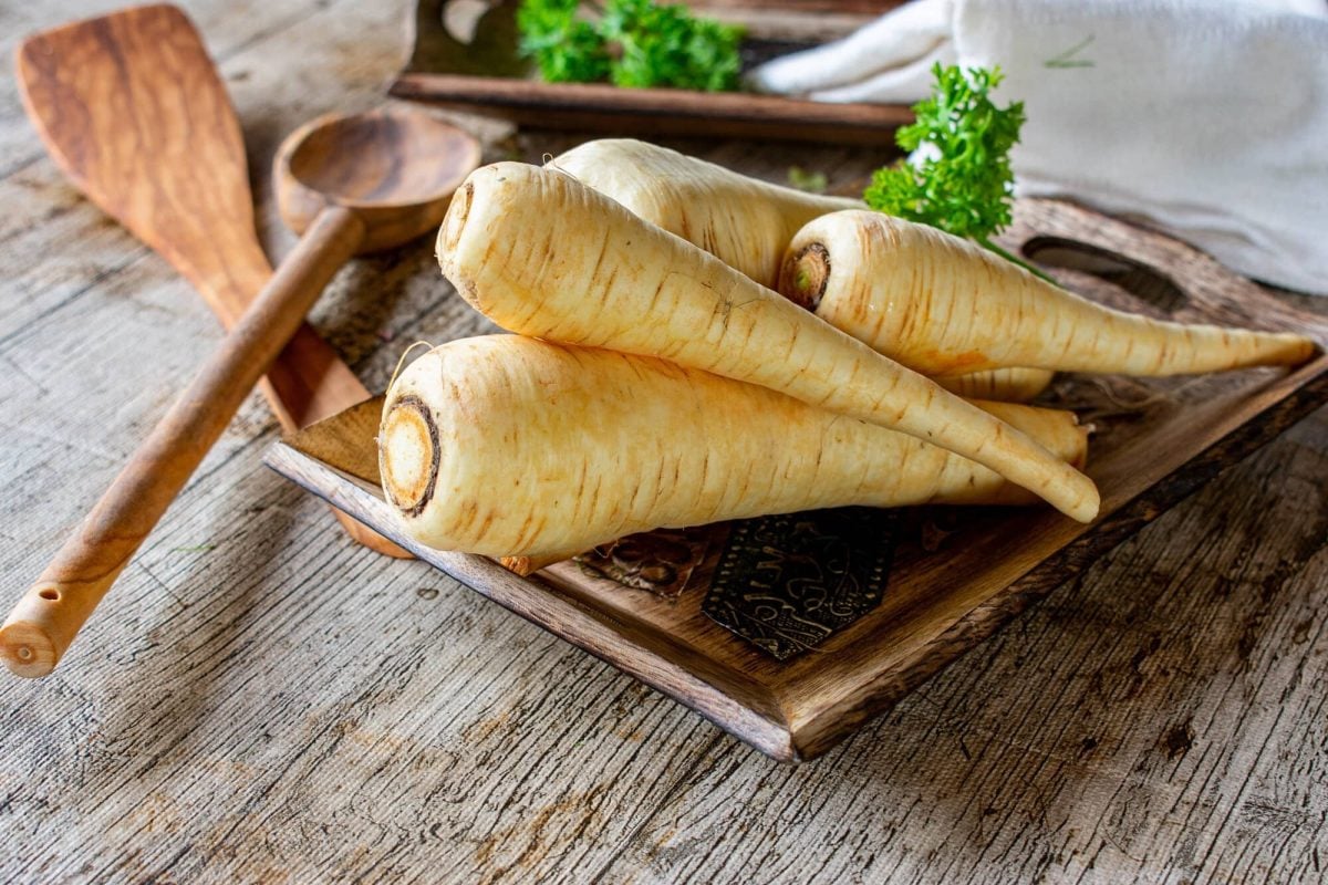 Frontalsicht: auf einem einer holzplatte liegen mehrere unbearbeitete rohe Pastinaken, frische Kräuter und zwei Kochlöffel aus Holz.