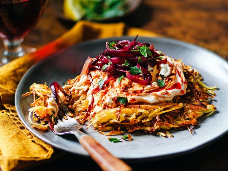 Schrägsicht: Brauner Untergrund auf dem im Bildzentrum ein grauer Teller mit einem Pastinakenrösti mit Harissa-Feta-Creme, karamellisierten Zwiebeln und frischer Petersilie zu sehen ist. Außerdem eine senfgelbe Stoffservierte und eine Gabel mit Holzgriff. Im Hintergrund ein Glas Rotwein.