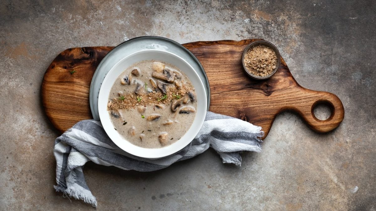Draufsicht: braun-gräulicher Steinuntergrund. In der Bildmitte ein Holzbrett mit einem Suppenteller, einer kleinen Schüssel und einem blau-weiß gestreiften Küchentuch. In der Schale Pilzcremesuppe mit Knoblauch und Thymian.