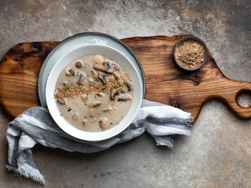 Draufsicht: braun-gräulicher Steinuntergrund. In der Bildmitte ein Holzbrett mit einem Suppenteller, einer kleinen Schüssel und einem blau-weiß gestreiften Küchentuch. In der Schale Pilzcremesuppe mit Knoblauch und Thymian.