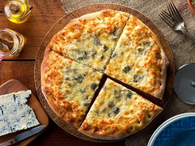 Draufsicht: eine Pizza Margherita mit Blauschimmelkäse die in 4 teile geschnitten wurde liegt auf einem Holztisch. Daneben liegt ein Blauschimmelkäse sowie ein Besteck.