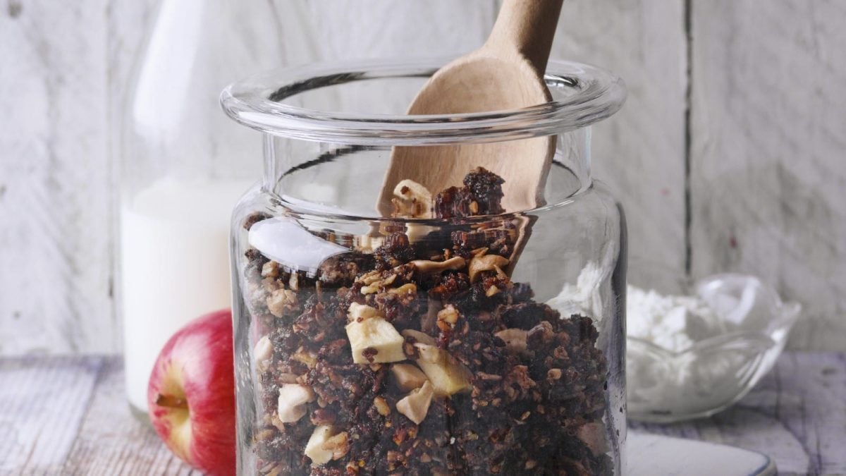Nahaufnahme eines Glases mit rheinischem Schwarzbrot Granola, in dem ein Holzlöffel steckt. Im Hintergrund ist außerdem eine Kanne Milch und ein Apfel zu sehen. Alles in einer hellen Holzumgebung.