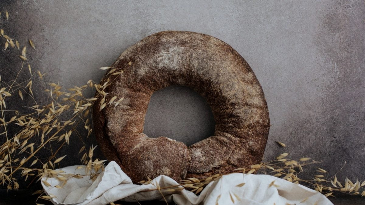 Graue Wand als Hintergrund. Daran lehnt ein Kringel Roggenbrot. Im Bildvordergrund ist außerdem eine weiße Leinenservierte und etwas Hafer zu sehen.