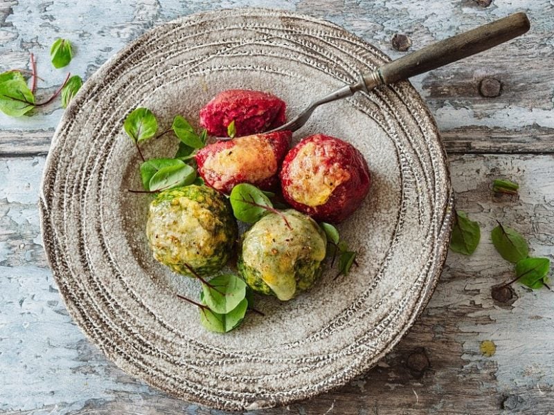 Rote-Bete-Knödel und Spinatknödel mit Parmesan überbacken auf einem rustikalen Teller, darauf und daneben Kräuter, vor Holzhintergrund.