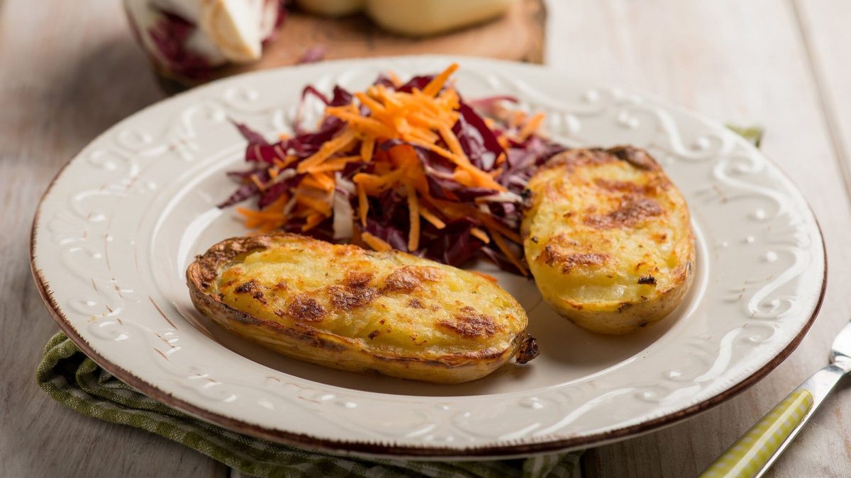 Schräge ansicht: Auf einem Holzuntergrund liegt ein Teller. Auf dem Teller liegen zwei Kartoffelhälften, die mit Käse überbacken wurden. Auch ein Rohkostsalat liegt auf dem teller. Und neben eine eine Gabel.