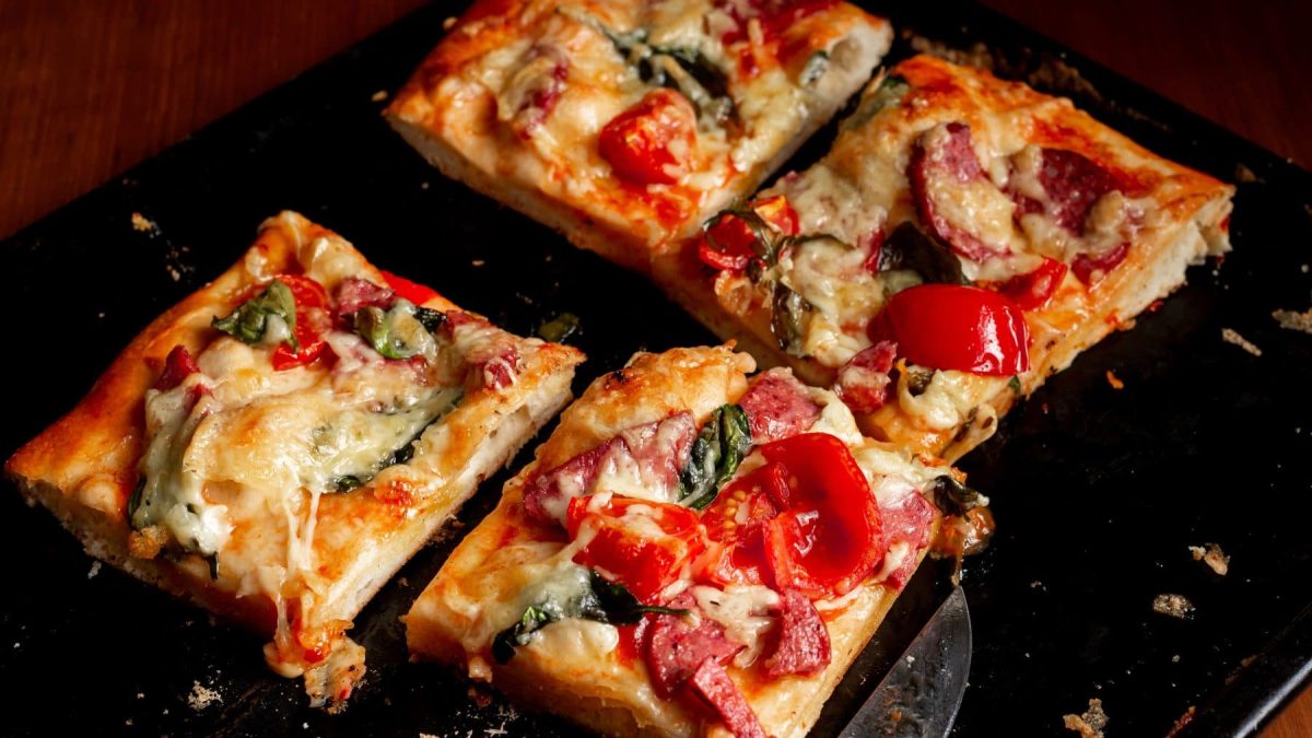 Skyr-Pizza ohne Hefe in 30 Minuten in vier Stücken mit Messer auf schwarzer Platte. Draufsicht.