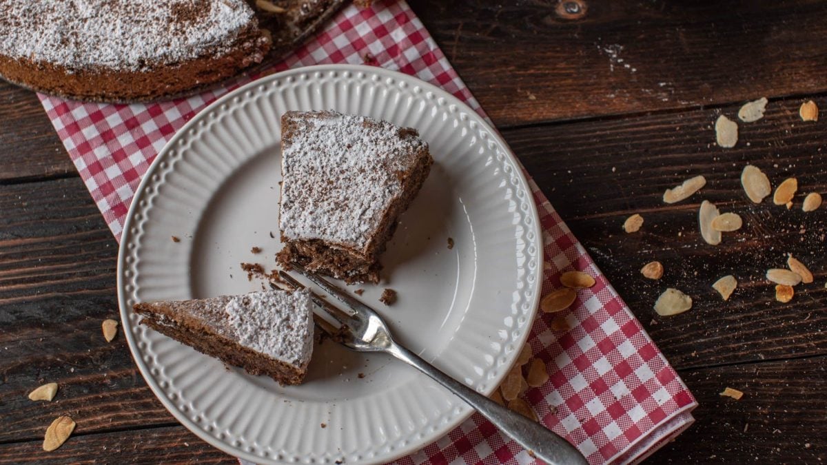 Torta Caprese al cioccolato Stück auf weißem Teller auf rot-weißem Tuch mit Kuchengabel. Daneben Mandelsplitter. Oben Torta Caprese al cioccolato im Ganzen. Draufsicht auf dunklem Holztisch.