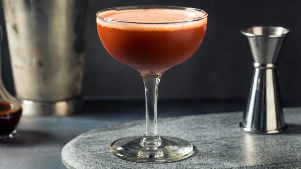 Trinidad Sour auf Steinplatte mit Barmaß. Im Hintergrund Shaker und eine Karaffe mit Mandelsirup. Frontalansicht.
