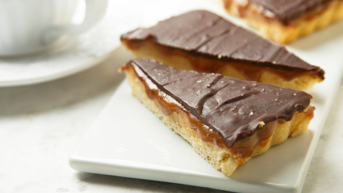 Karamell Cookies wie Twix Twix Cookies auf weißem Teller in Dreiecksform. Im HIntergrund eine Tasse mit Untersetzer. Draufsicht.