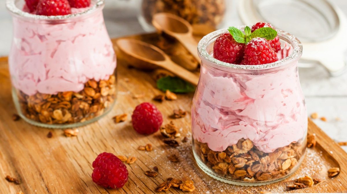 Zwei Gläser mit Himbeer-Mousse und Müsli, dekoriert mit frischen Himbeeren, Minze und Puderzucker. Darum liegen Himbeeren und Müsli.