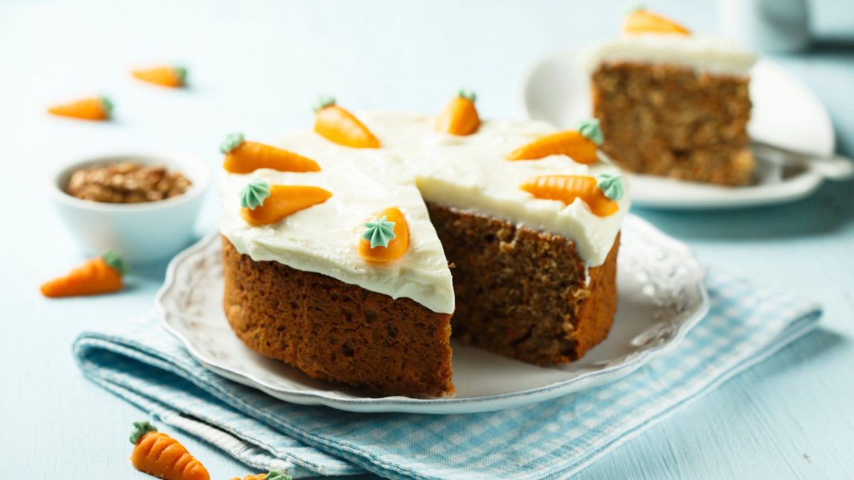 Ein angeschnittener veganer Schoko-Karottenkuchen mit weißem Frosting auf einem Teller, dahinter steht ein weiterer Teller mit einem Stück Kuchen.