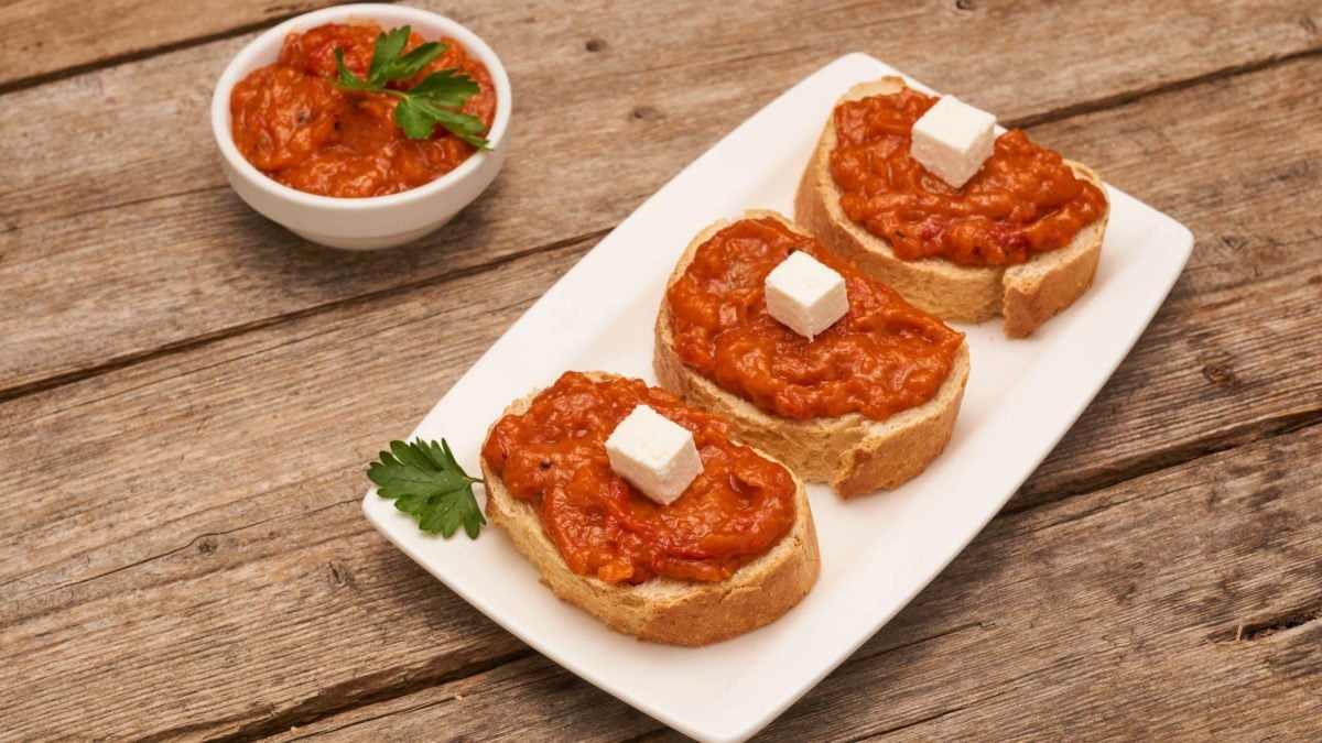 Ajvar-Feta-Creme mit Feta-Würfel auf drei Scheiben Brot auf weißem Teller und Holz. Daneben Schale mit der Creme. Draufsicht.