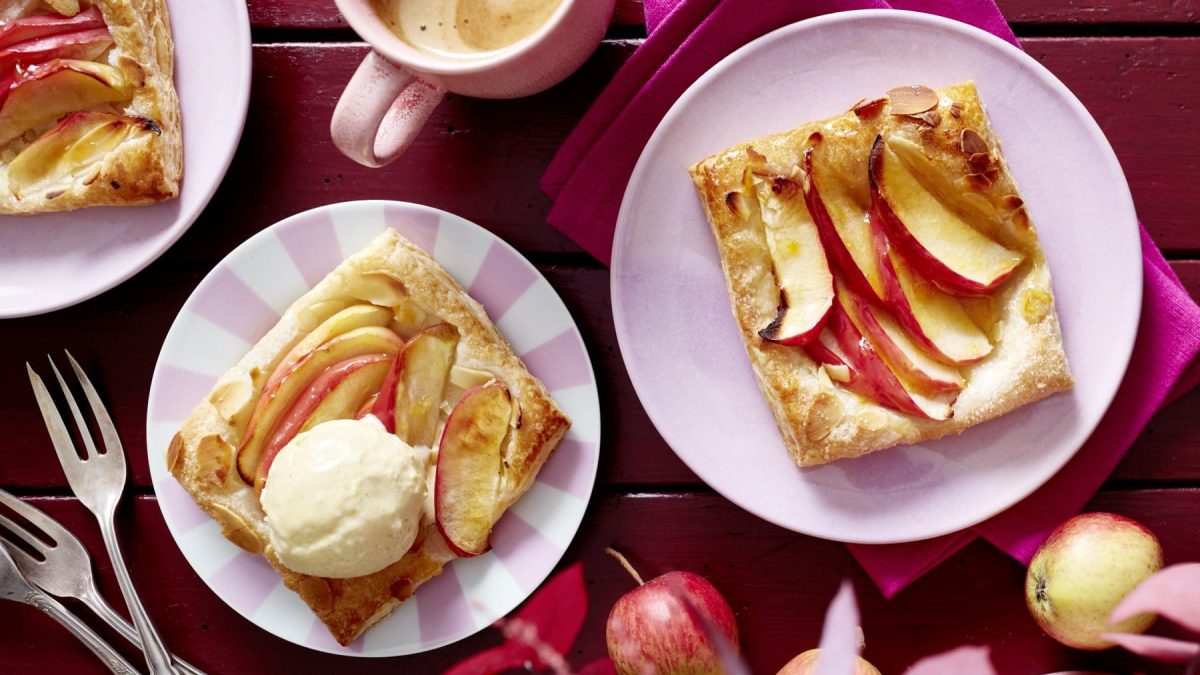 Apfel-Knusper-Teilchen mit Vanilleeis auf rosa Tellern vor Holzhintergrund in der Draufsicht.
