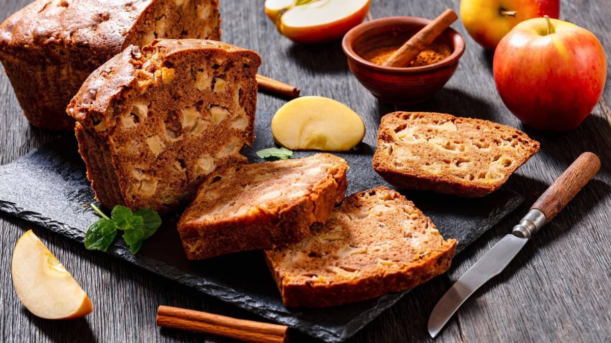 Draufsicht: Ein Apfel-Zimt-Brot liegt aufgeschnitten mit Äpfeln und Zimt auf einem dunkleren Holztisch.