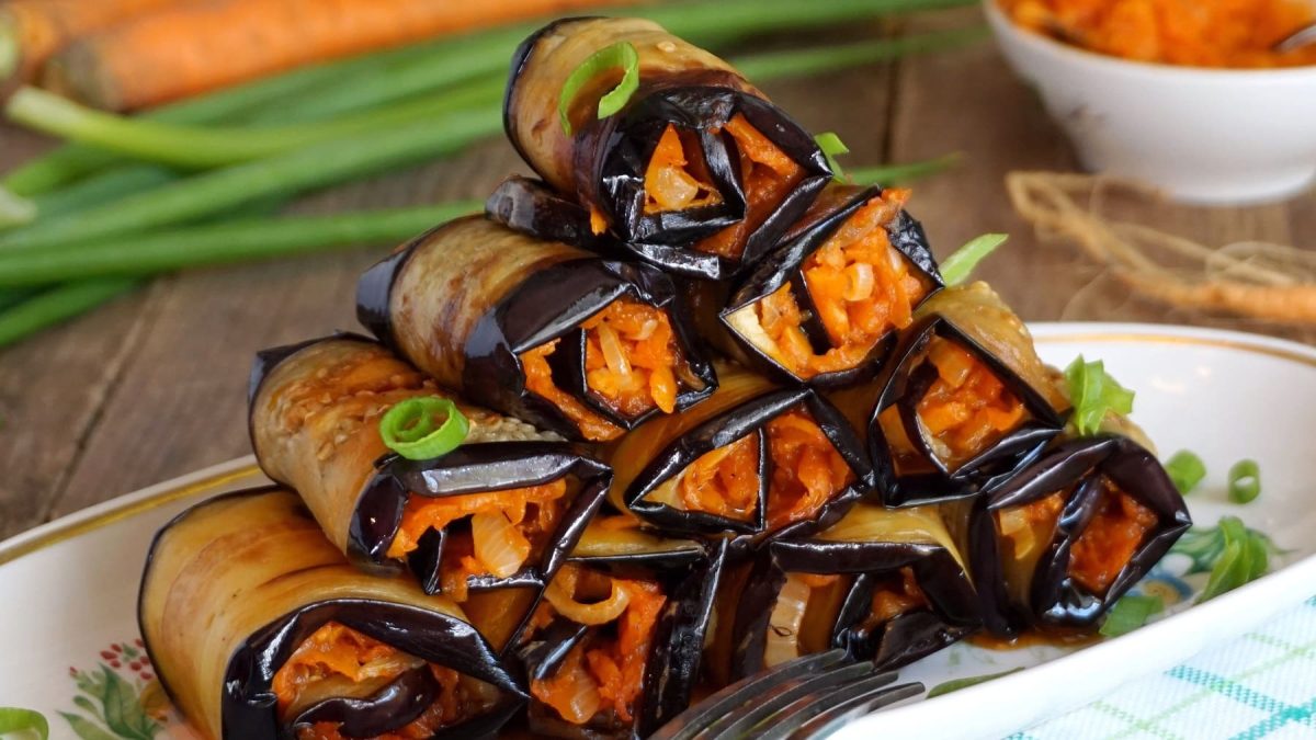 Mehrere Auberginen-Röllchen mit Kimchi und Karotten gefüllt auf einem länglichen weißen Teller.